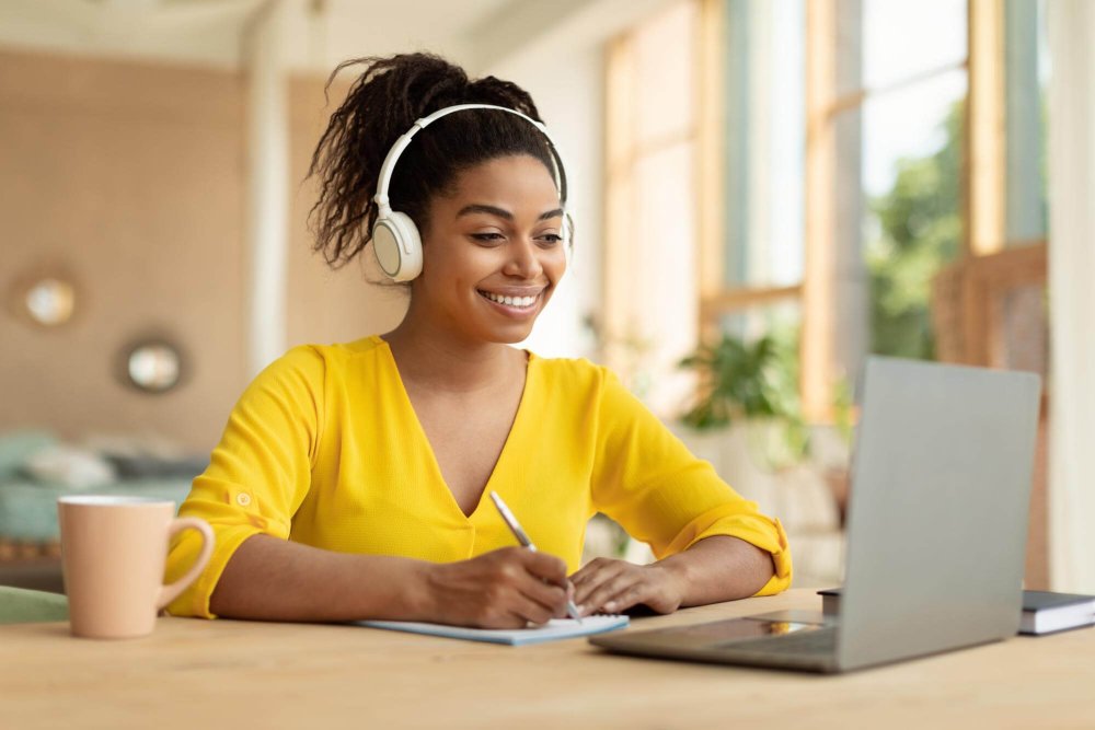smiling-black-lady-wireless-headphones-working-laptop-taking-notes-paper-notebook-watching-webinar